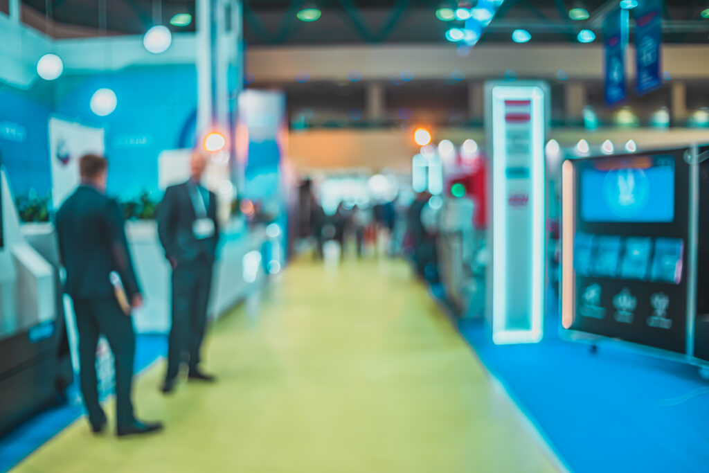 A blurred image of a corporate trade show in an indoor setting.