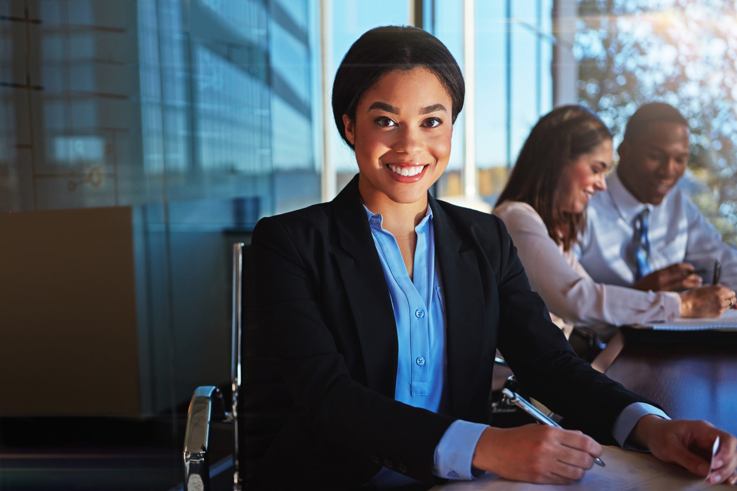 A young business professional in a bright, modern office, working on pen and paper with her colleagues discussing in the background.
